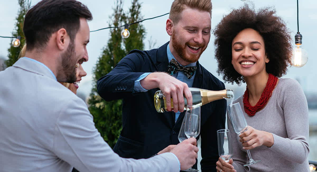 People smiling and laughing while pouring a glass of white wine into wine flutes. 