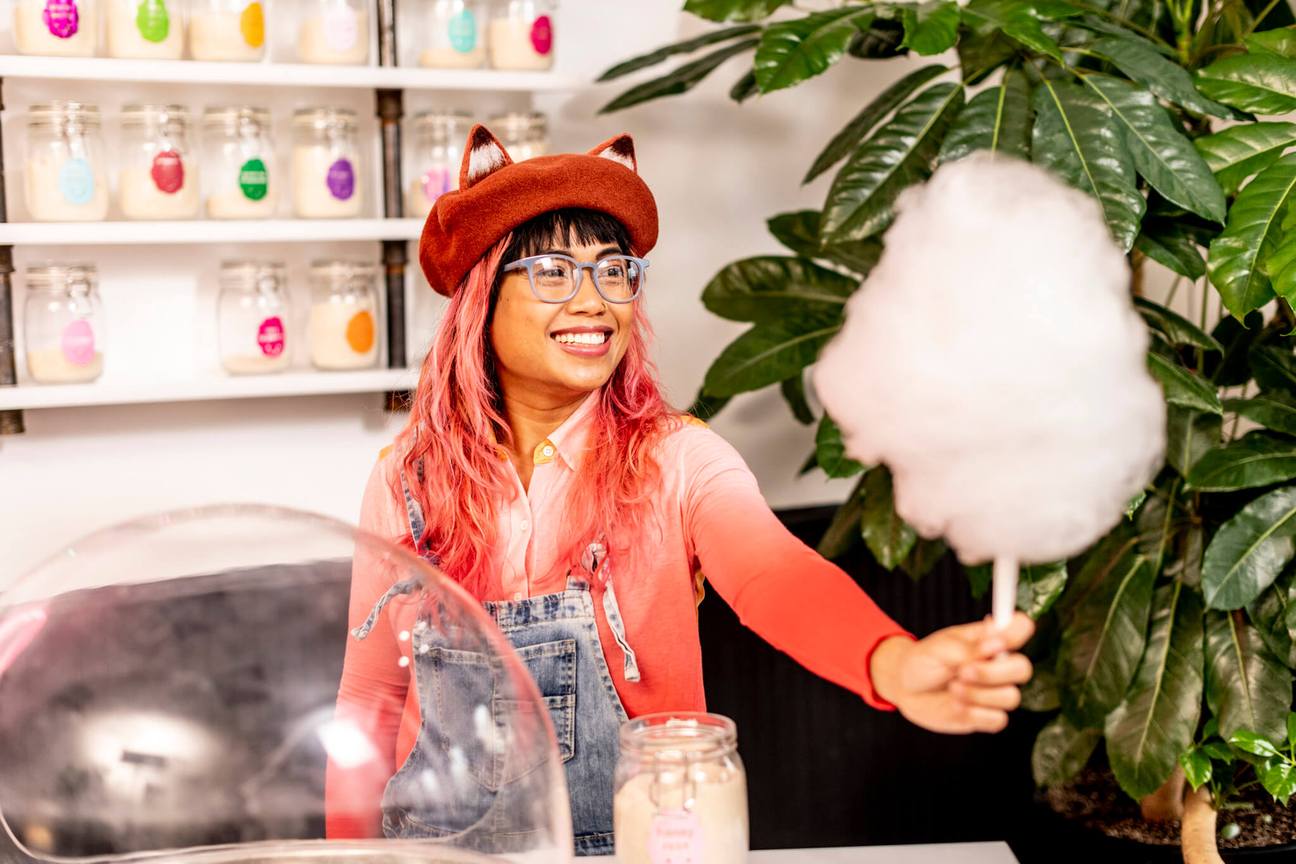 Smiling person with pink hair, a red beret, and silver-framed spectacles, holding out a fluffy and generous-sized portion of cotton candy. 