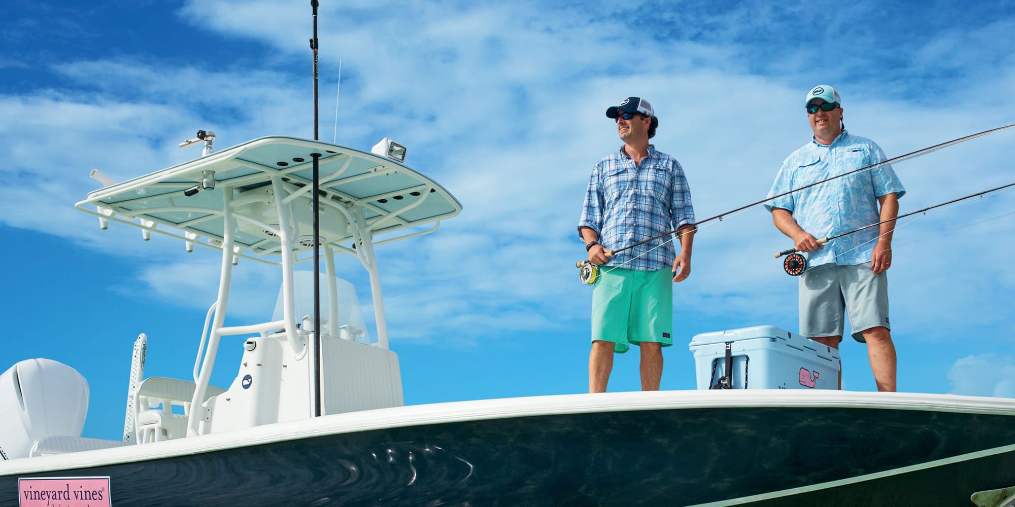 Two men on boat holding fishing poles near a cooler with a vineyard vines sticker.