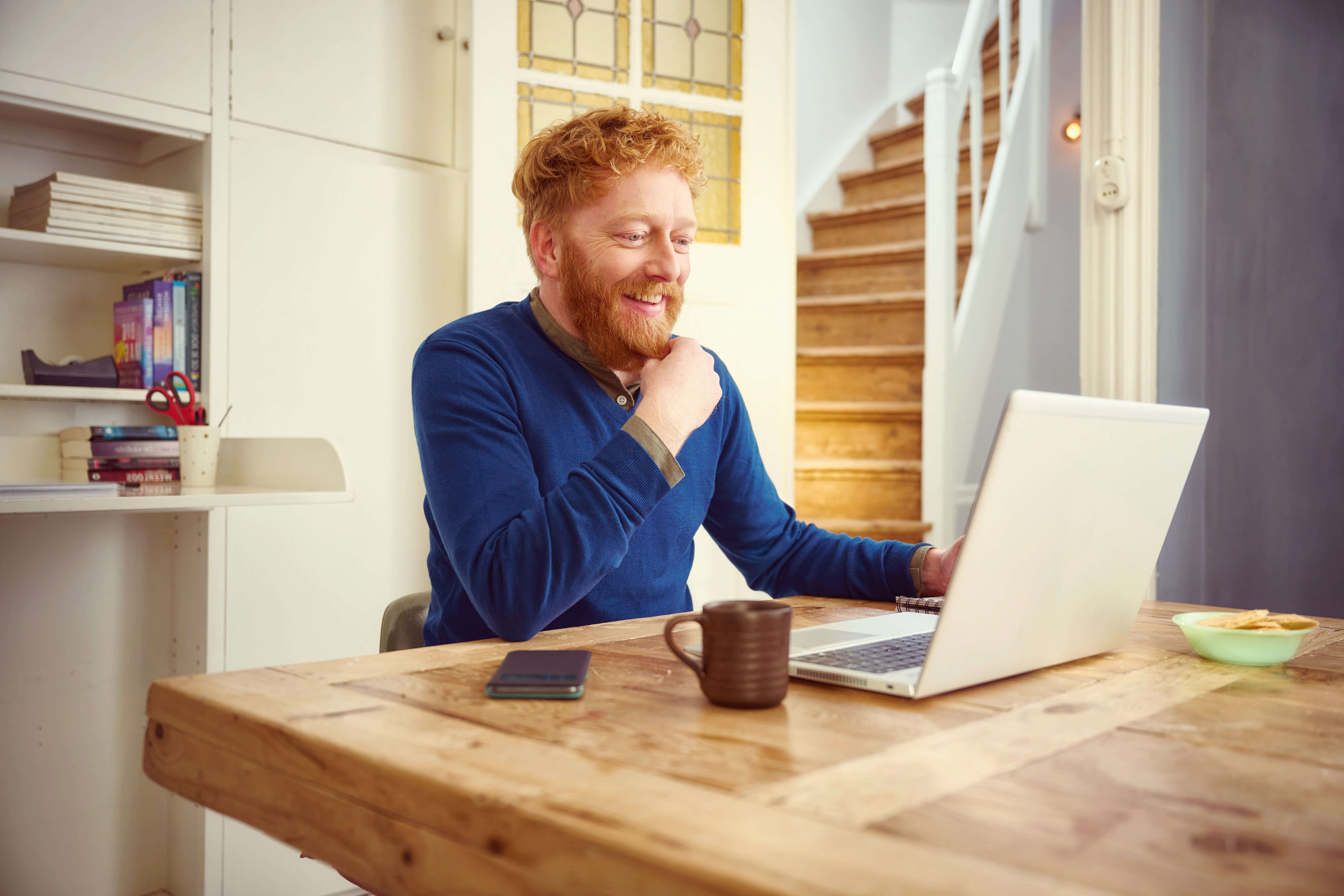 Een lachende man zit achter zijn laptop met een bruine koffiemok en een zwarte telefoon op tafel naast zich.