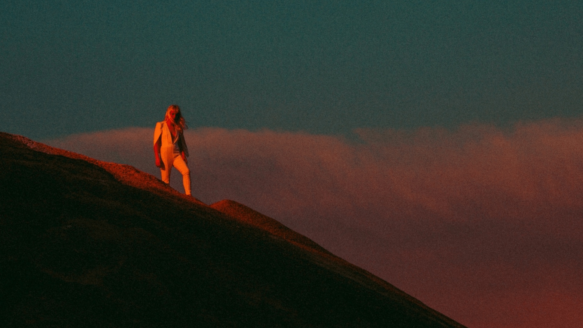 Person with mid length blonde hair standing on top of hill with grainy blue sky and red hues from sundown