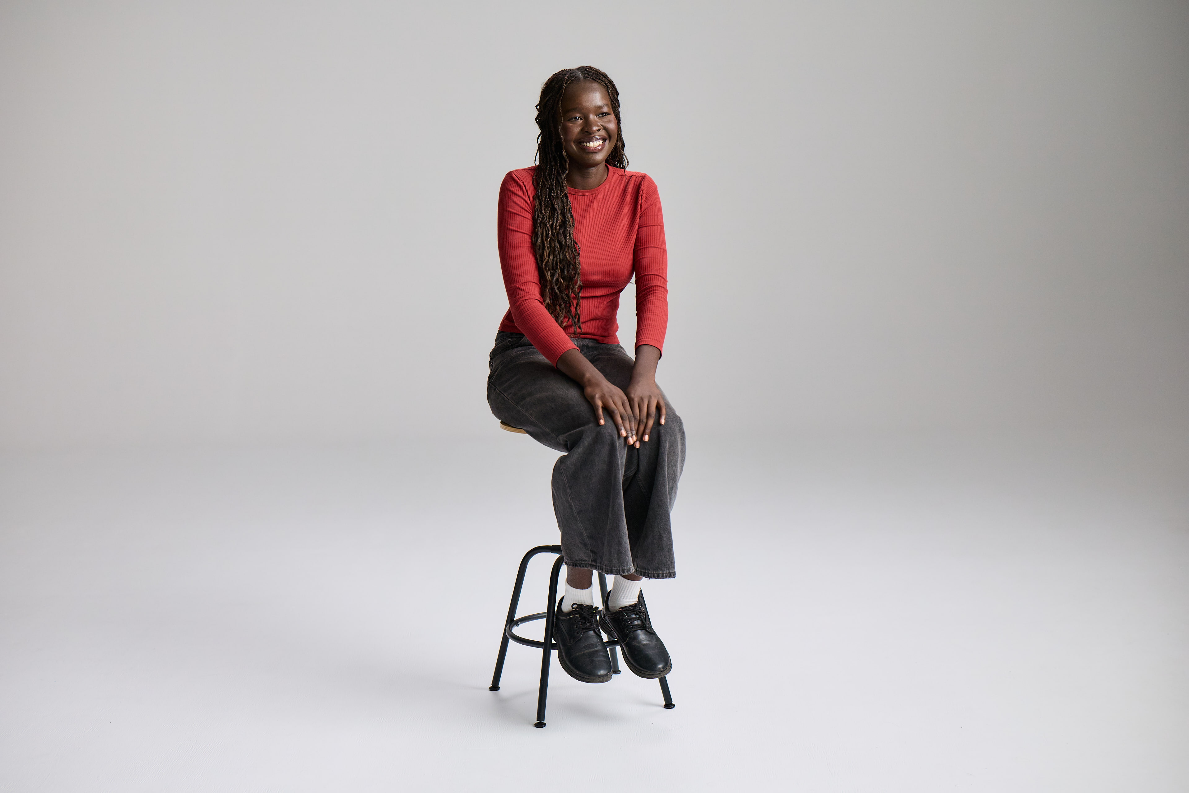 Full-body studio portrait of a young woman with long, dark braids, sitting on a black stool. She is smiling and wearing a red long-sleeved shirt and grey jeans. This photo is for a Red Cross Lifeblood campaign.