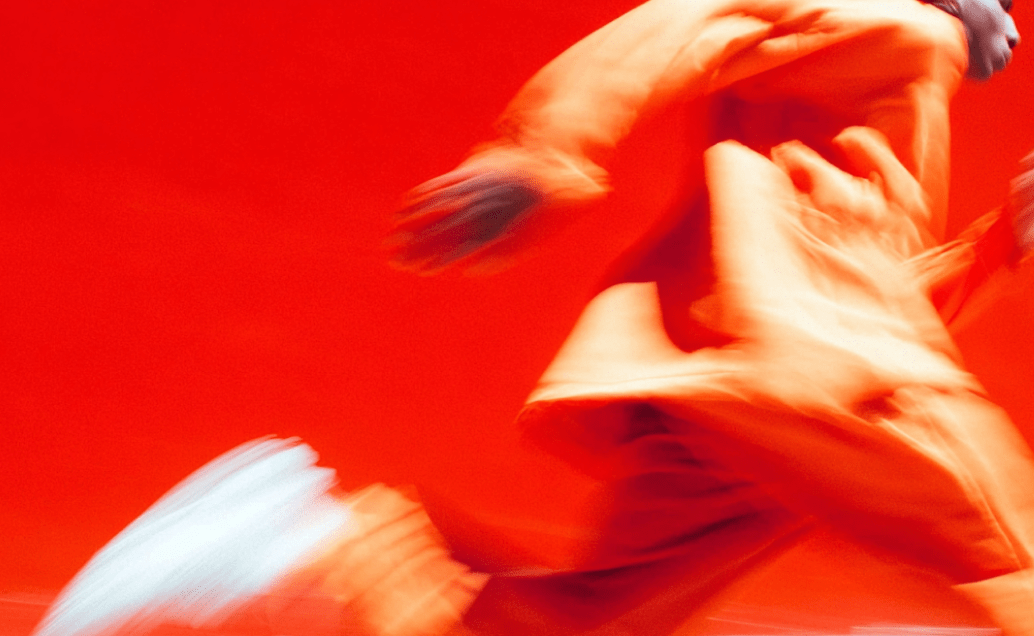 Abstract, motion-blurred image of a person running against a vibrant red background, wearing an orange outfit. A bright white sneaker blur is visible.