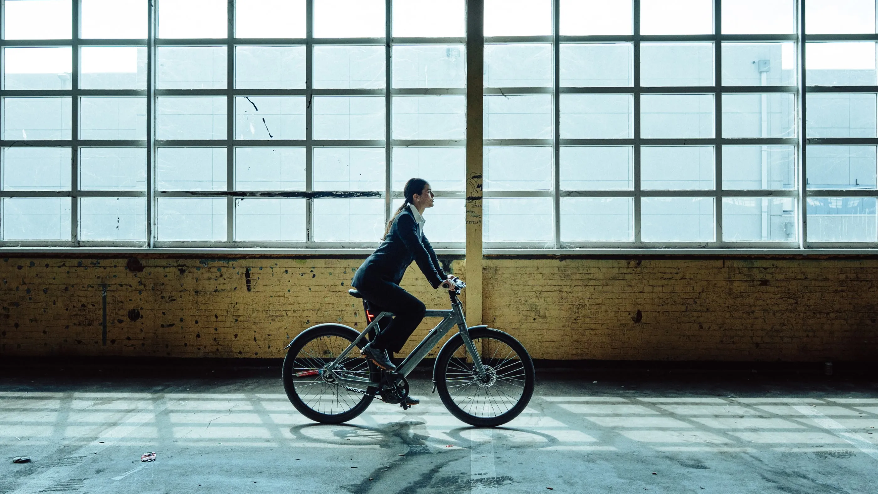 Woman in business attire riding a bicycle inside a warehouse with large industrial windows.
