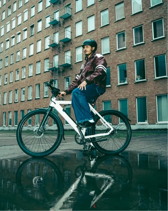 Man on a white modern bicycle reflected in a puddle in front of a brick apartment building.