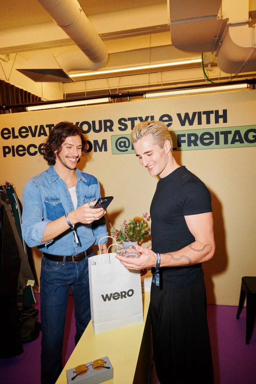 Two people stand at a display table in a stylish store, smiling while looking at a phone and a small item. A WERO shopping bag and accessories are on the table behind them.