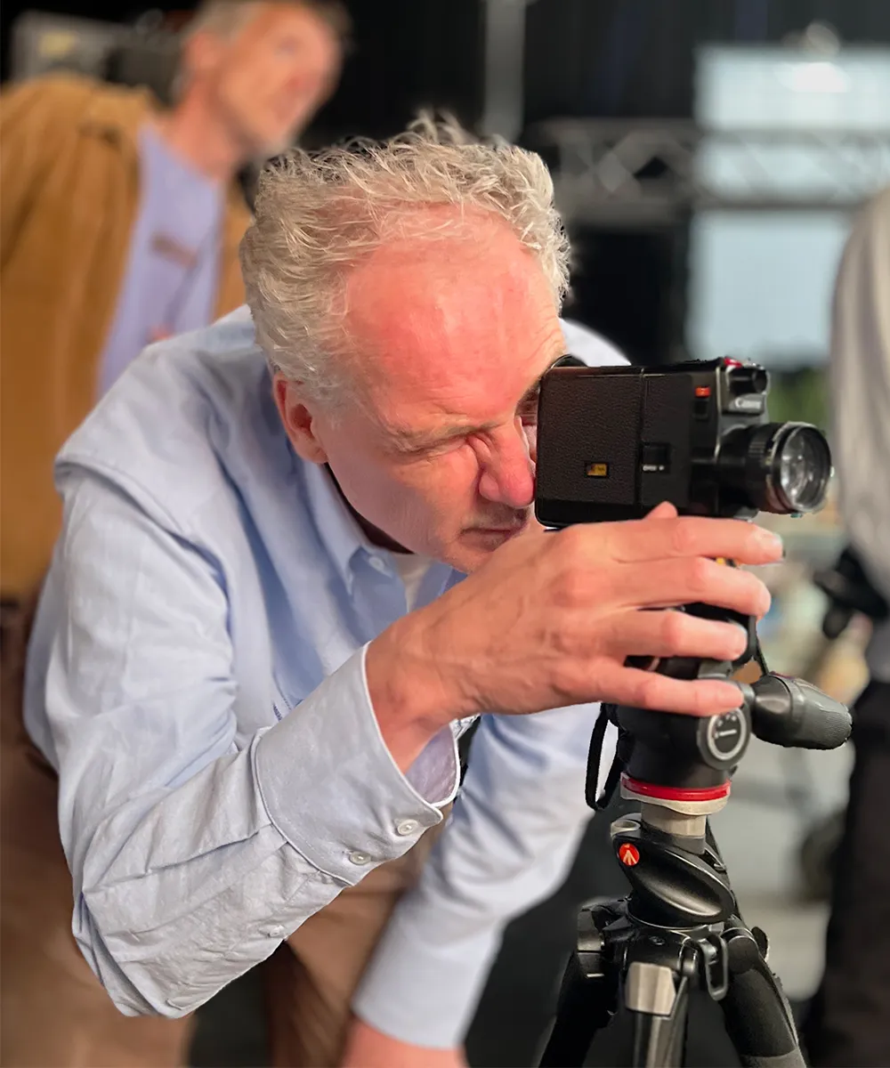 Film director looking intensely through the viewfinder of a classic film camera on a tripod.