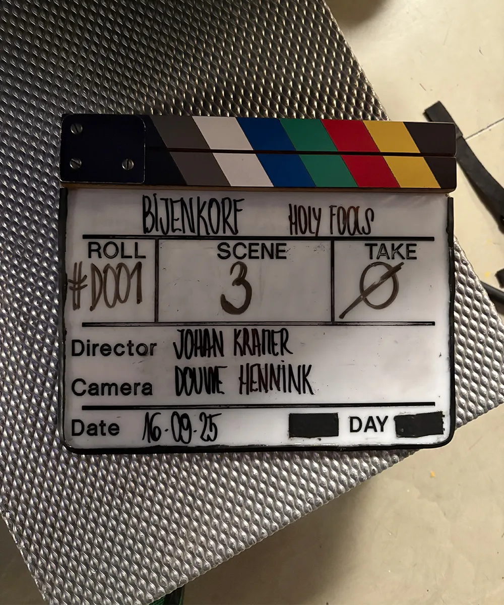 A film clapperboard for a production titled 'BIJENKORF HOLY FOOLS' with scene details.