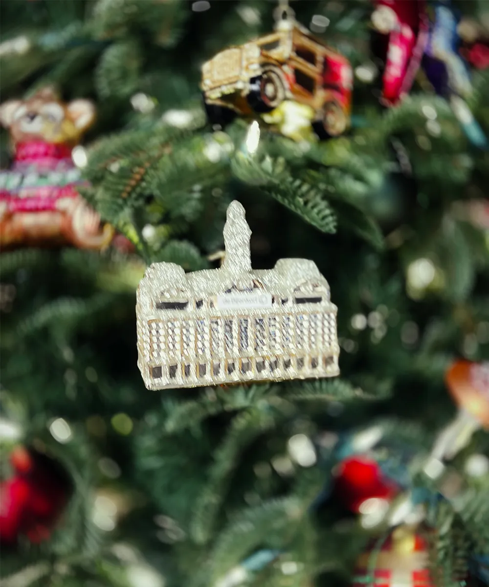 A glittery Christmas ornament shaped like the De Bijenkorf flagship store building, hanging on a Christmas tree.