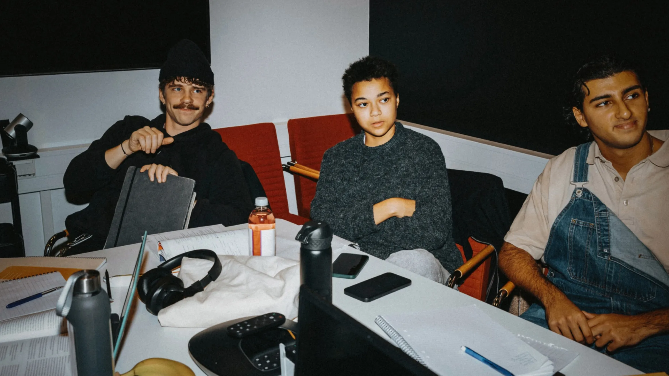 A photo of three university students sitting around a table with study materials, laptops, and water bottles. The student on the left wears a black beanie and a mustache; the student in the middle looks engaged in a discussion; and the student on the right is wearing denim overalls over a beige polo shirt.